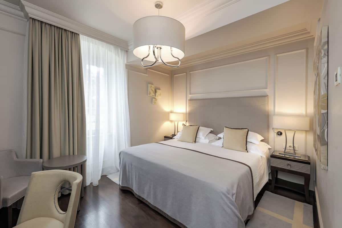 Ein Hotelzimmer mit Bett, Nachttischen, einem Sessel, einer Lampe und einem großen Fenster mit Vorhängen.