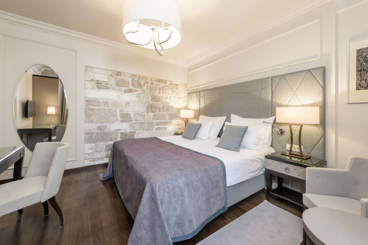 Ein Hotelzimmer mit einer Wand aus Naturstein, einem großen Bett mit grauem Kopfteil, zwei Nachttischen mit Lampen und einem runden Spiegel.