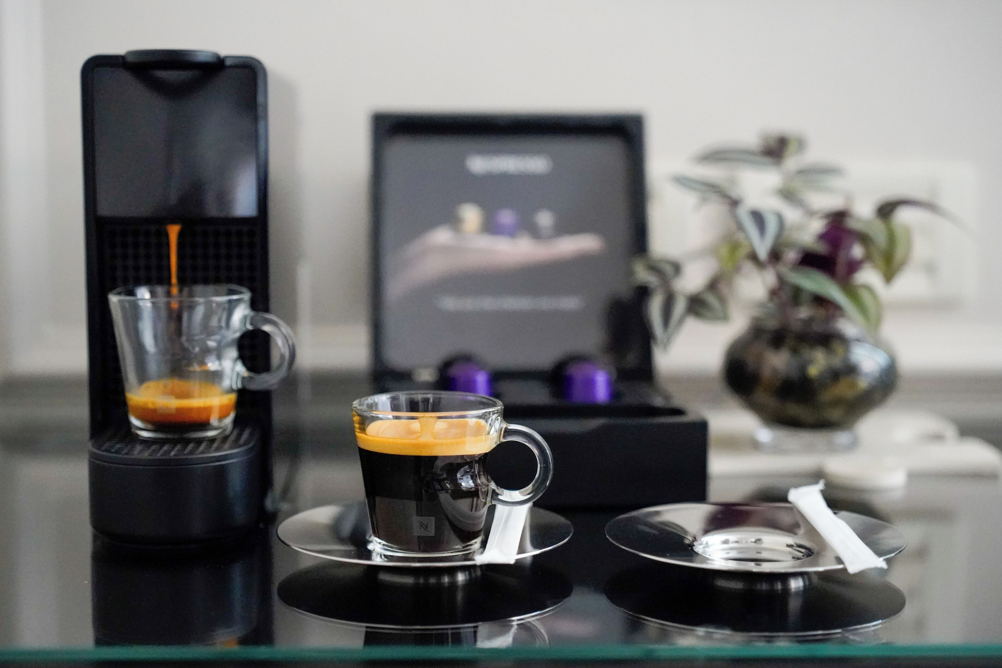 A coffee machine dispensing coffee into a glass cup on a glass table.