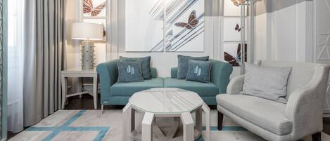 A modern living room with a teal sofa, a grey armchair, a glass-top coffee table, and a white side table with a lamp.