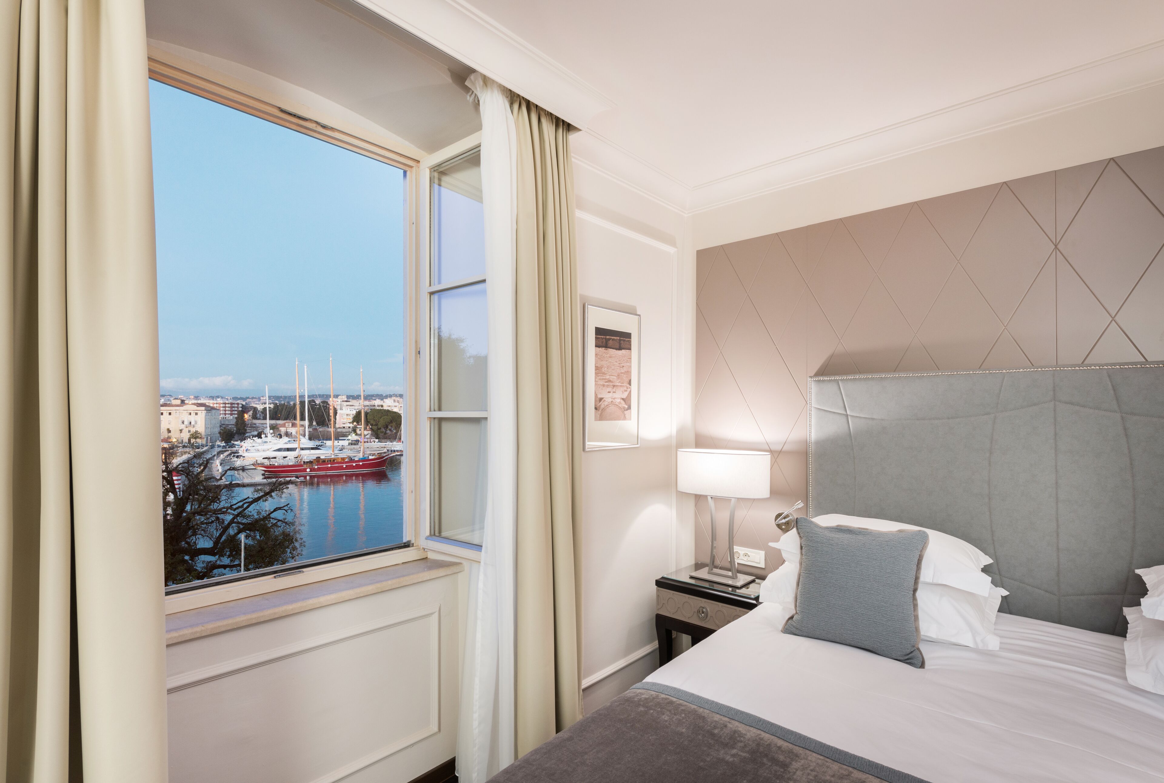 A hotel room with a bed, a nightstand, a lamp, a window with curtains, and a view of a marina.