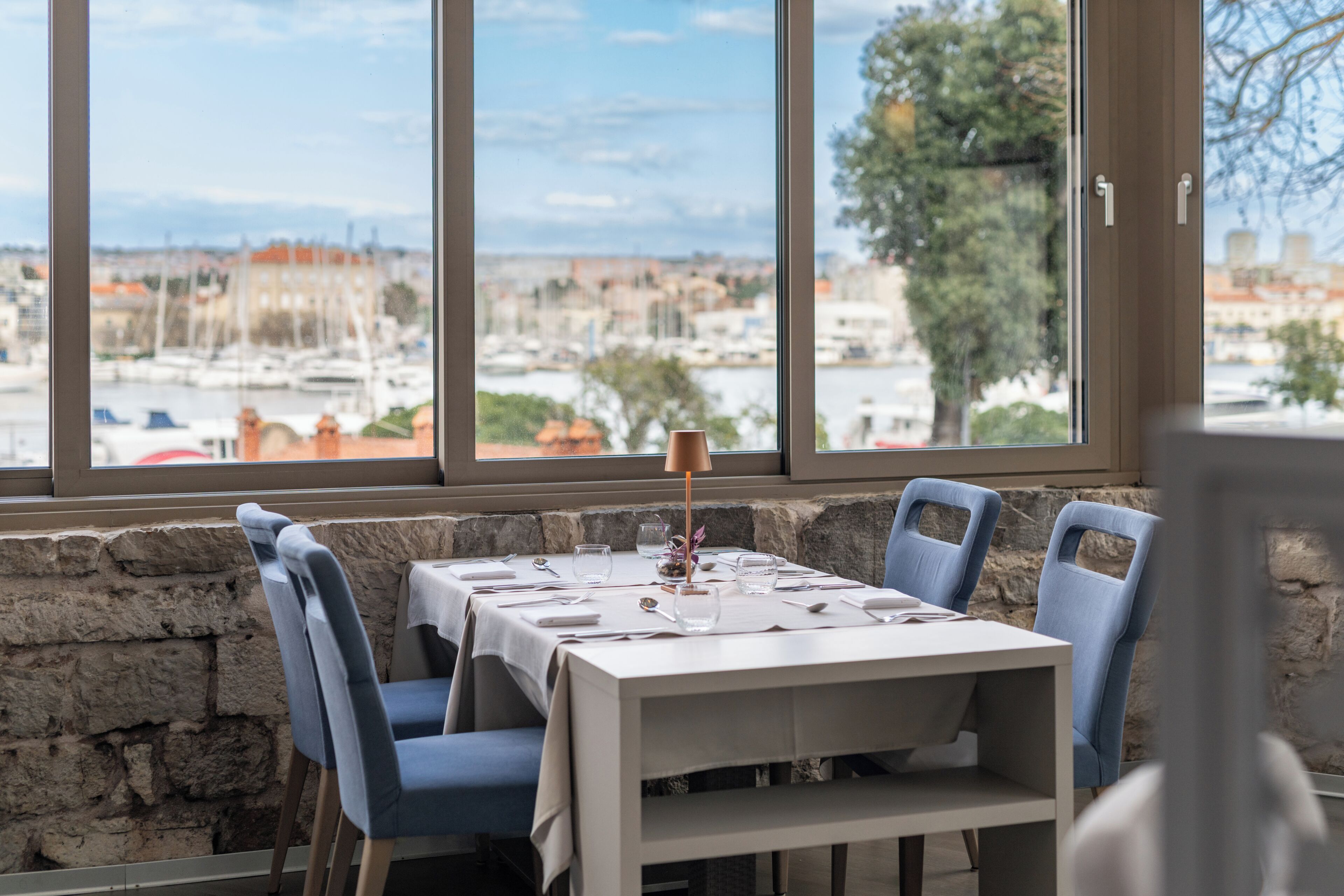 A dining table with chairs, a lamp, and a view of a marina and buildings through large windows.