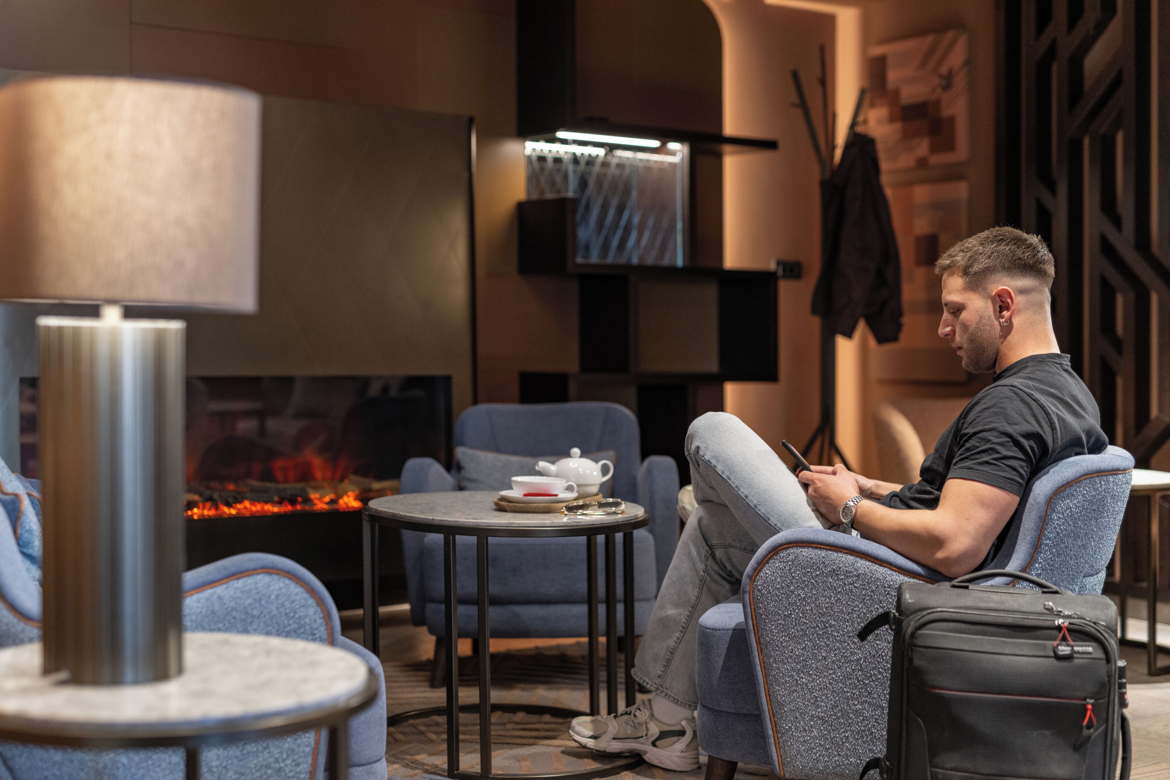 A man sitting on a chair in a modern room with a fireplace, a table with a teapot, and a small table with a lamp.