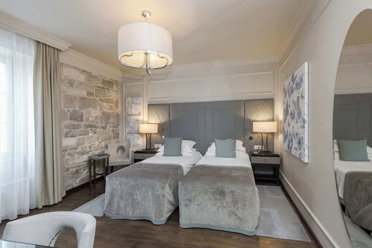 Ein Hotelzimmer mit einer Wand aus Stein, zwei Betten mit grauer Bettwäsche, einer Kristallleuchtre und einem großen Kunstwerk an der Wand.