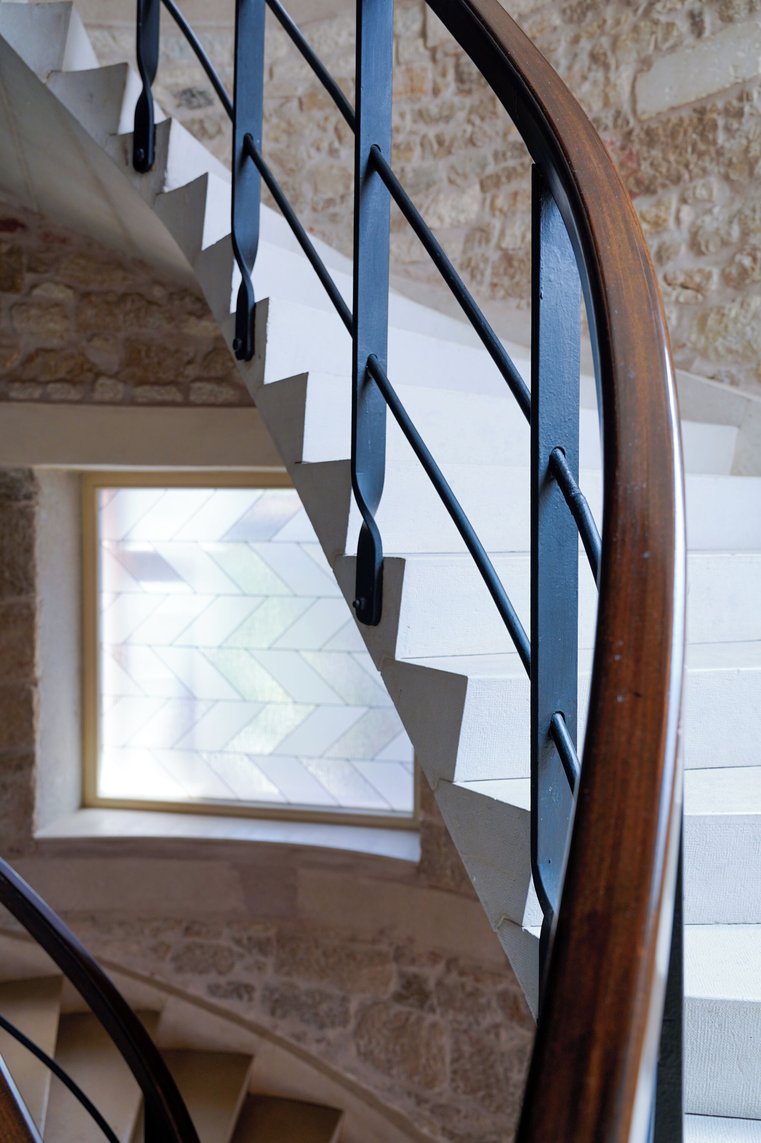 Un escalier en colimaçon, doté d’une rampe en bois et de balustrades en métal, mène à une pièce avec une fenêtre.