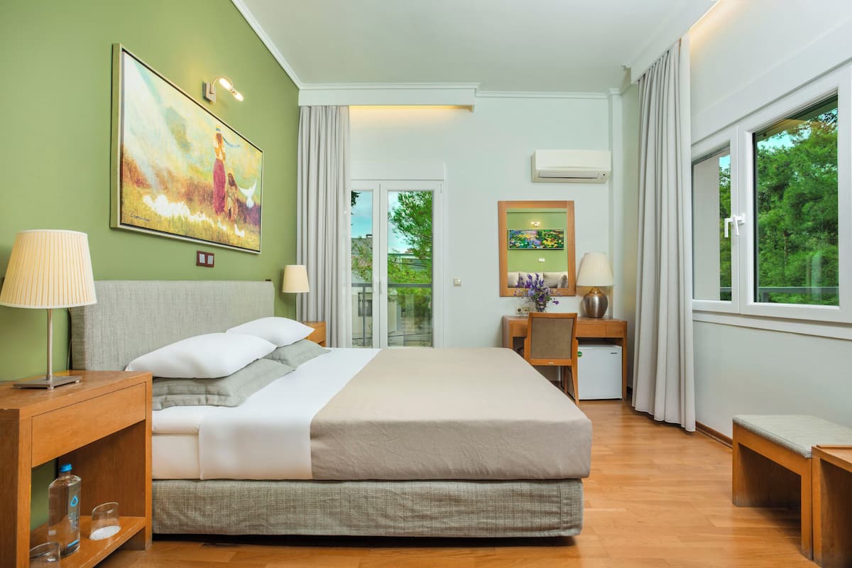A modern hotel room with a large bed, a desk, and a view of greenery.