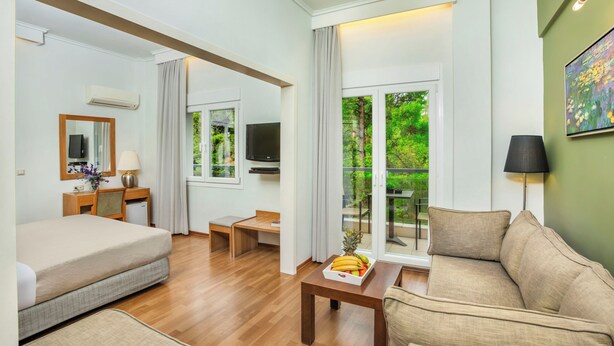 A modern hotel room with a bed, a sofa, a dining table with fruit, a TV, and a balcony with a view of greenery. - Pefka Hotel (Pylaia-Chortiatis)
