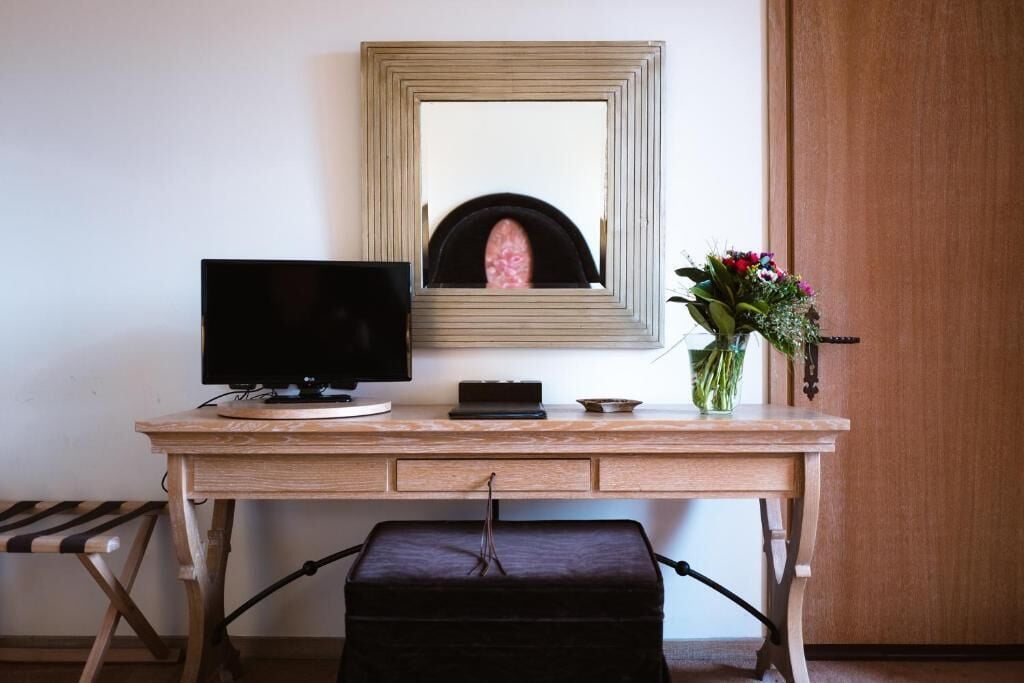 A wooden desk with a flat-screen TV, a mirror, a vase with flowers, and a black ottoman.