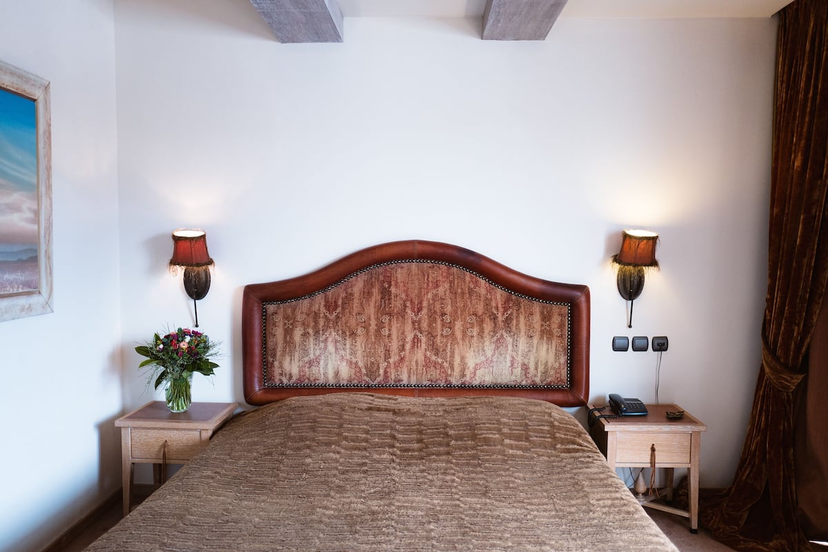 Ein Schlafzimmer mit einem hölzernen Kopfstück, einem Bett, Nachttischen mit Lampen, einer Blumenvase und einem Bild an der Wand.