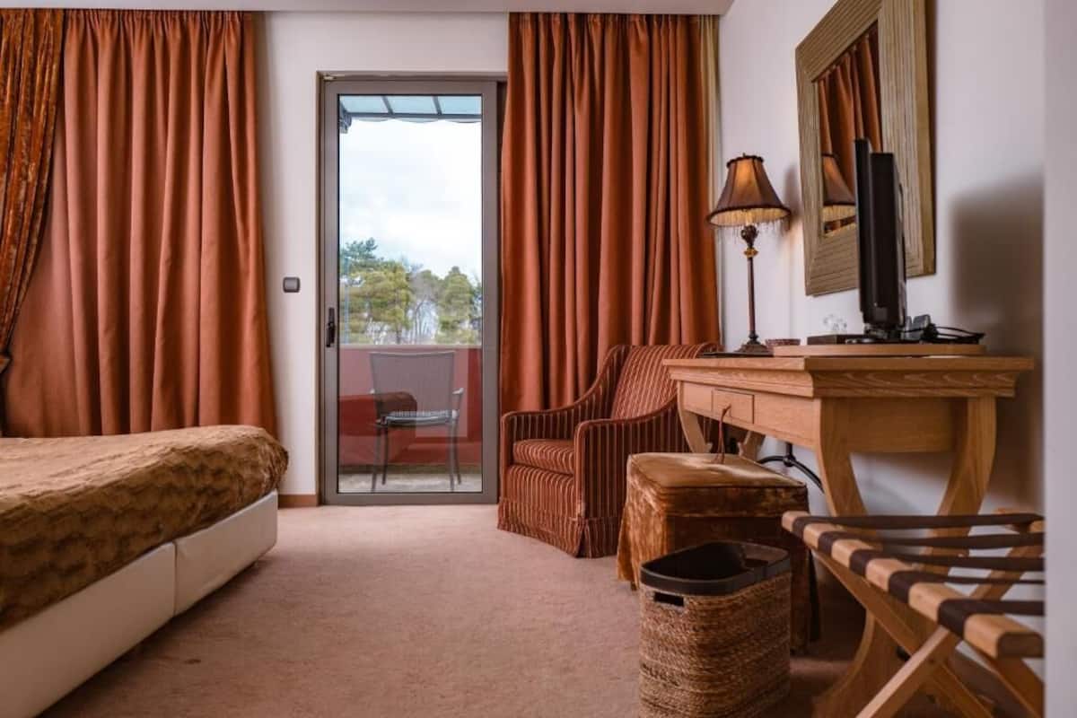 Ein Hotelzimmer mit Bett, Schreibtisch mit Lampe, Stuhl, kleinem Tisch, Papierkorb und einer Glastür zum Außenbereich.