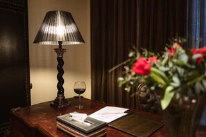 Un escritorio de madera con una lámpara, una copa de vino tinto, papeles y una jarra con flores rojas.