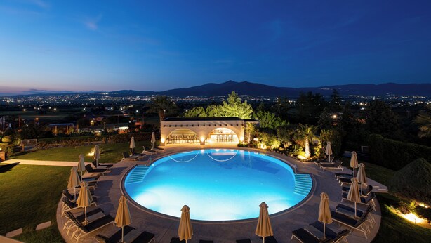 A large, illuminated swimming pool surrounded by lounge chairs and umbrellas, with a view of distant mountains and city lights. - Heaven Hotel (Thermi)