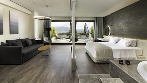 A modern hotel room with a large bed, a sofa, a dining table, and a balcony with a view of the mountains. - Heaven Hotel (Thermi)