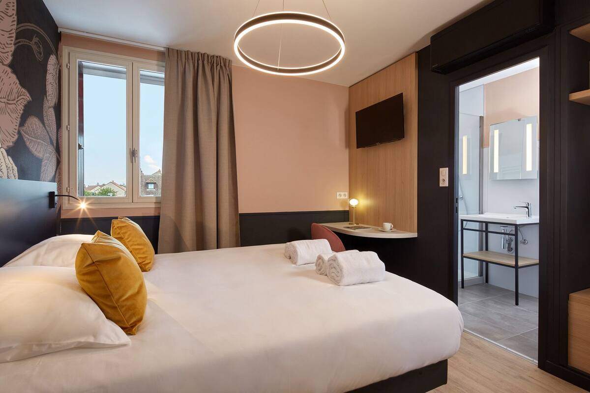 Une chambre d’hôtel avec un lit, un bureau, un miroir et une salle de bain visible à travers une porte ouverte.