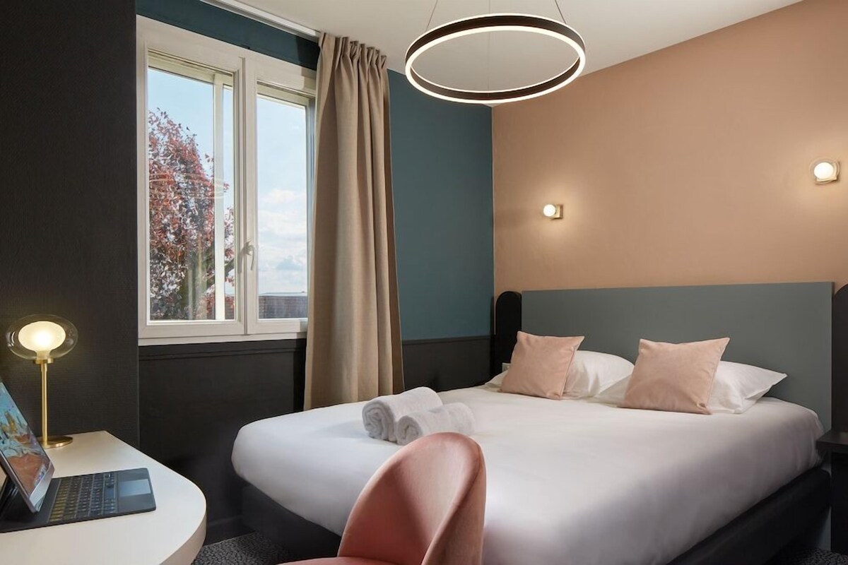 A hotel room with a bed, a desk with a laptop, a chair, and a window with curtains.