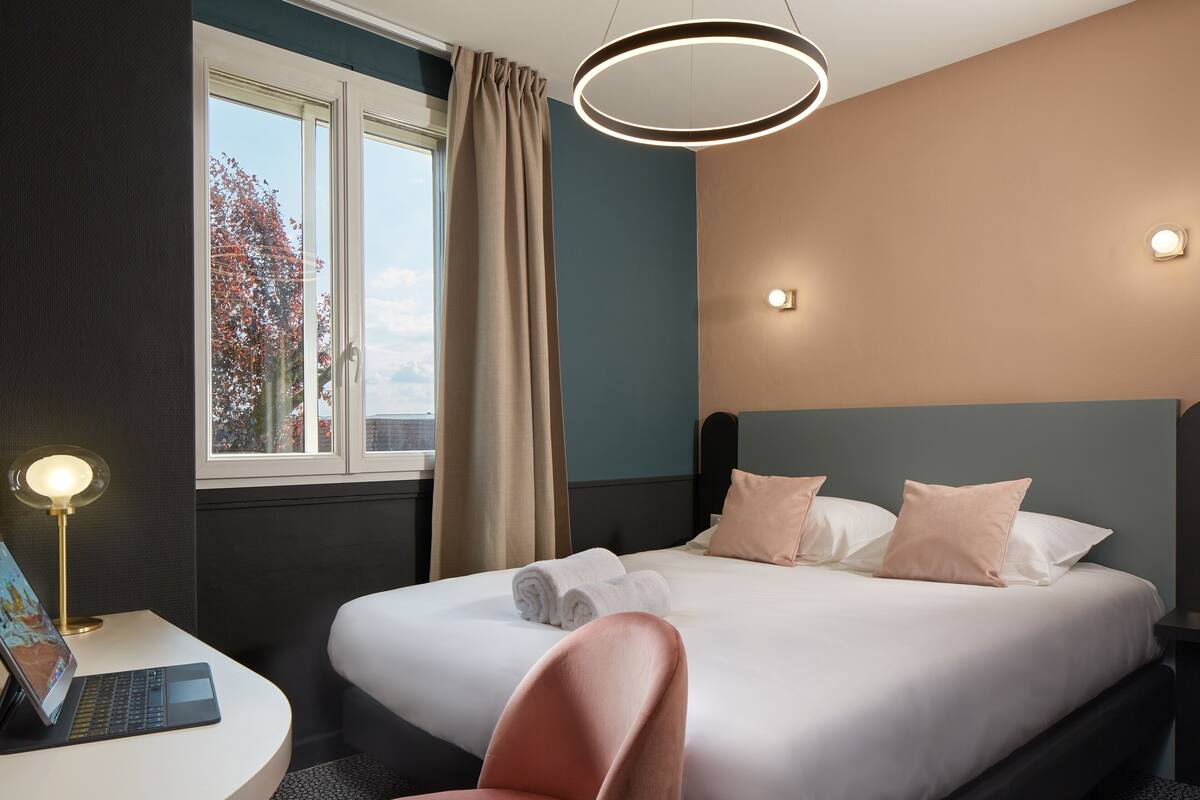Une chambre d’hôtel comprenant un lit, un bureau avec un ordinateur portable, une chaise, une fenêtre avec des rideaux et un luminaire fixé au mur.