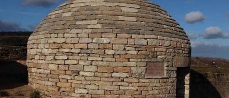 A stone structure with a rounded dome shape, possibly a historical building or monument, situated in a grassy area with a clear sky.
