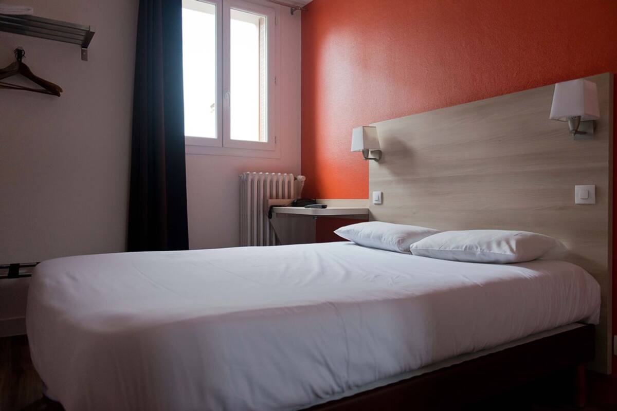 A hotel room with a bed, a bedside table, a lamp, a radiator, and a window with curtains.