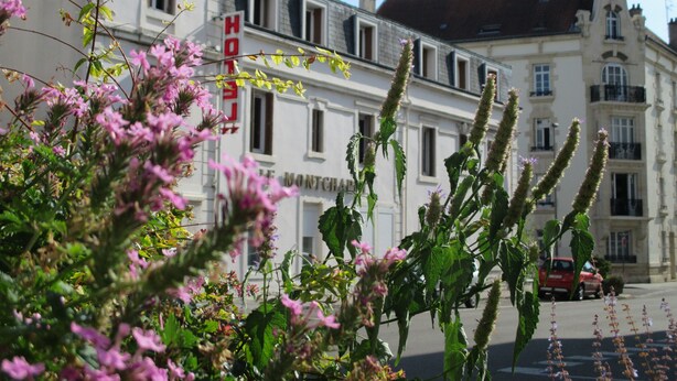 Un immeuble doté de nombreuses fenêtres et de balcons, une enseigne rouge avec la lettre « H », et une rue bordée de voitures garées.