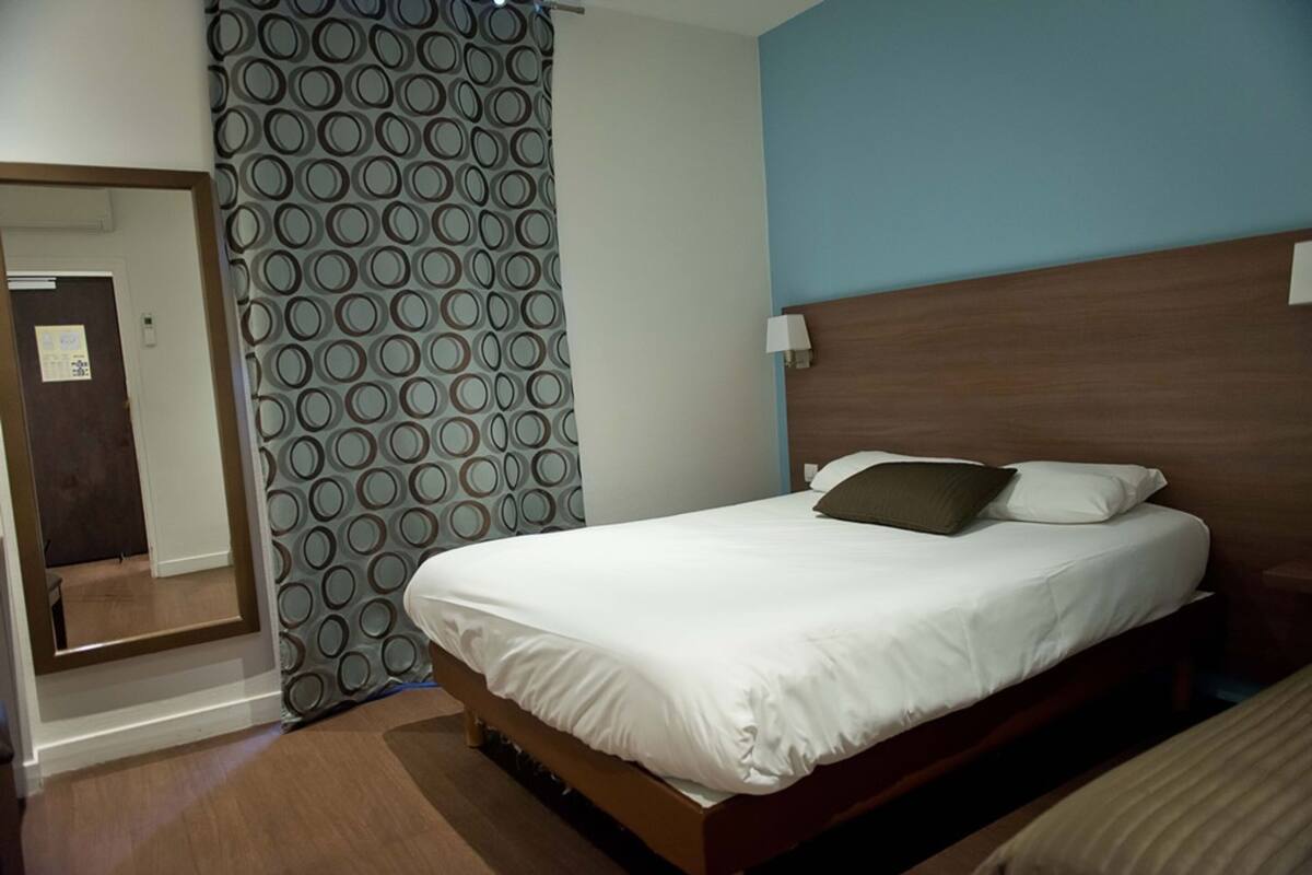 A hotel room with a bed, a mirror, a patterned curtain, and a nightstand with a lamp.