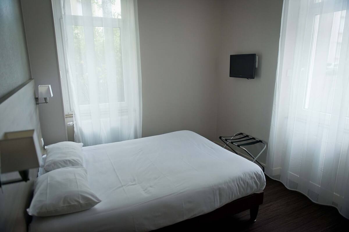 A neatly made bed with white linens, a bedside table with a lamp, and a TV mounted on the wall.