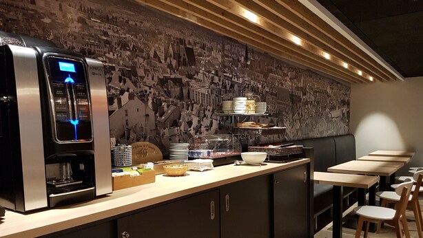 Un café moderno con una máquina de café, una barra con alimentos y una pared decorada con un mural de un paisaje urbano.