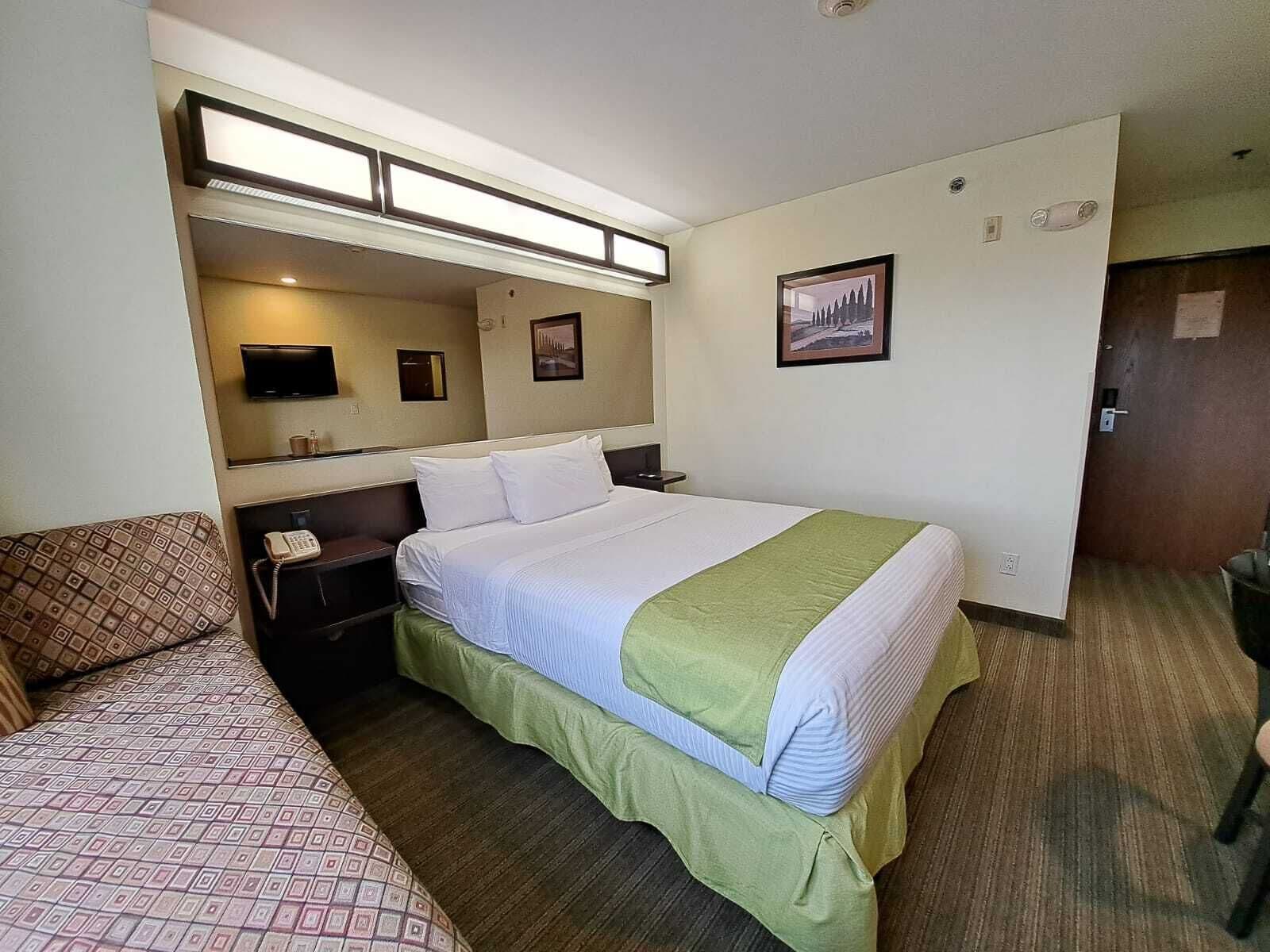 A hotel room with a large bed, a bedside table with a phone, a small table with a lamp, a couch, and a wall-mounted TV.