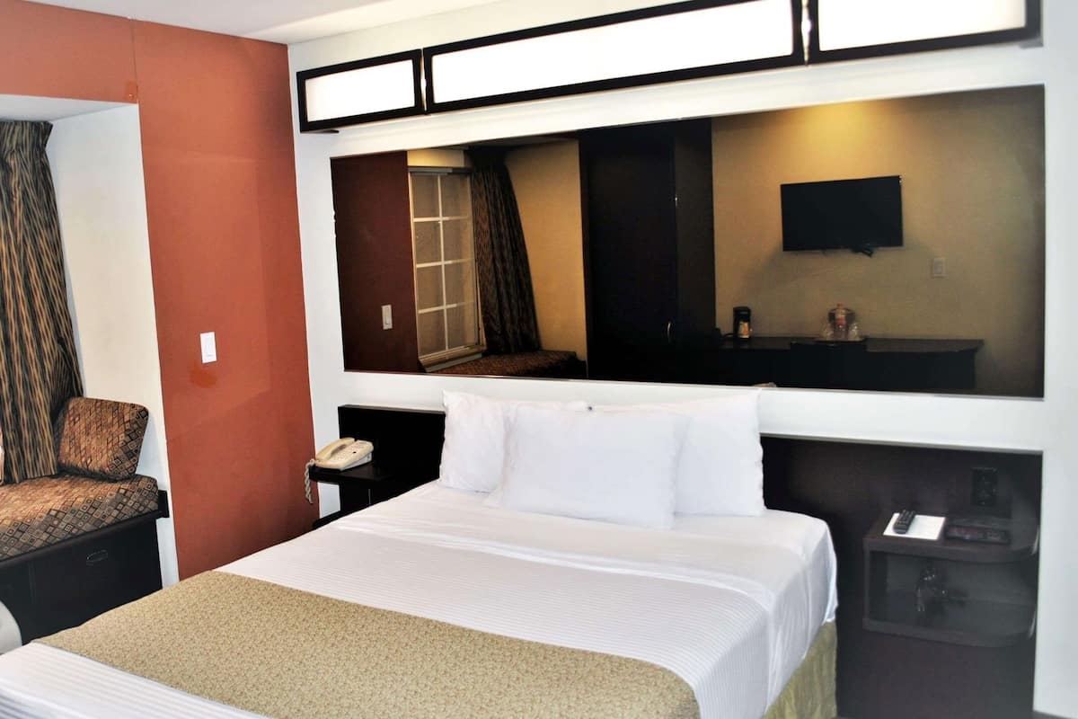 Habitación de hotel con cama, una silla, escritorio con teléfono, espejo y televisor.