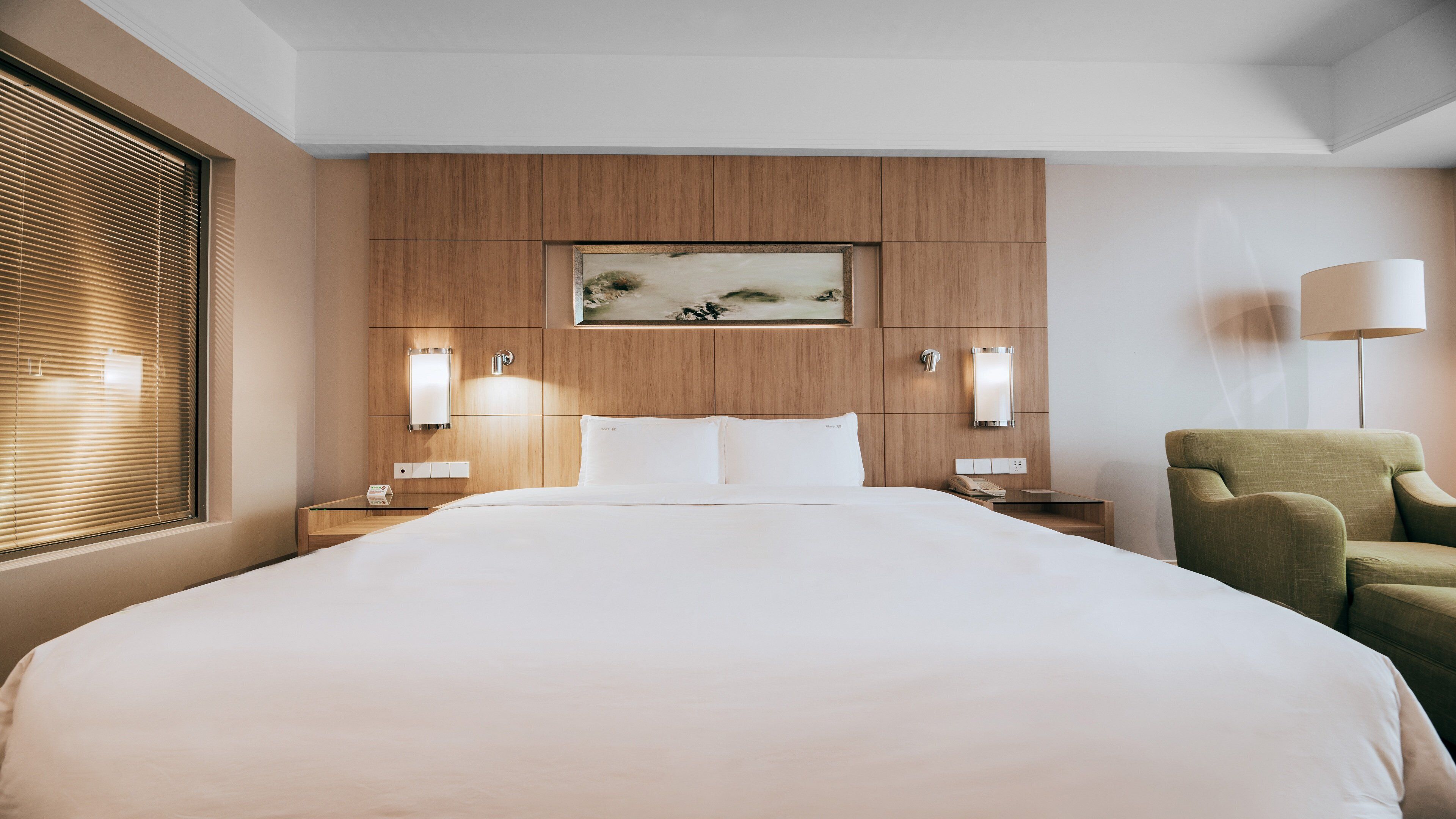 A hotel room with a large bed, wooden headboard, two bedside tables with lamps, a green armchair, and a painting above the bed.