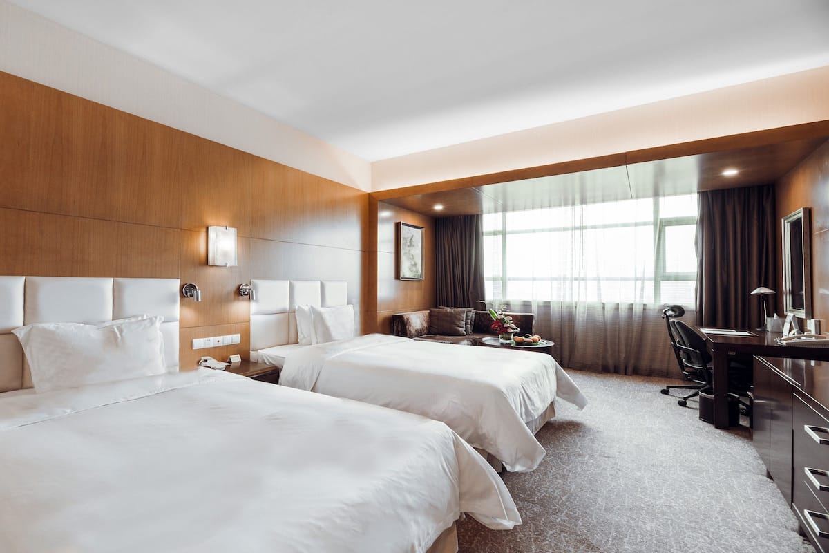 A hotel room with two beds, a desk, a chair, a desk lamp, and a large window with curtains.