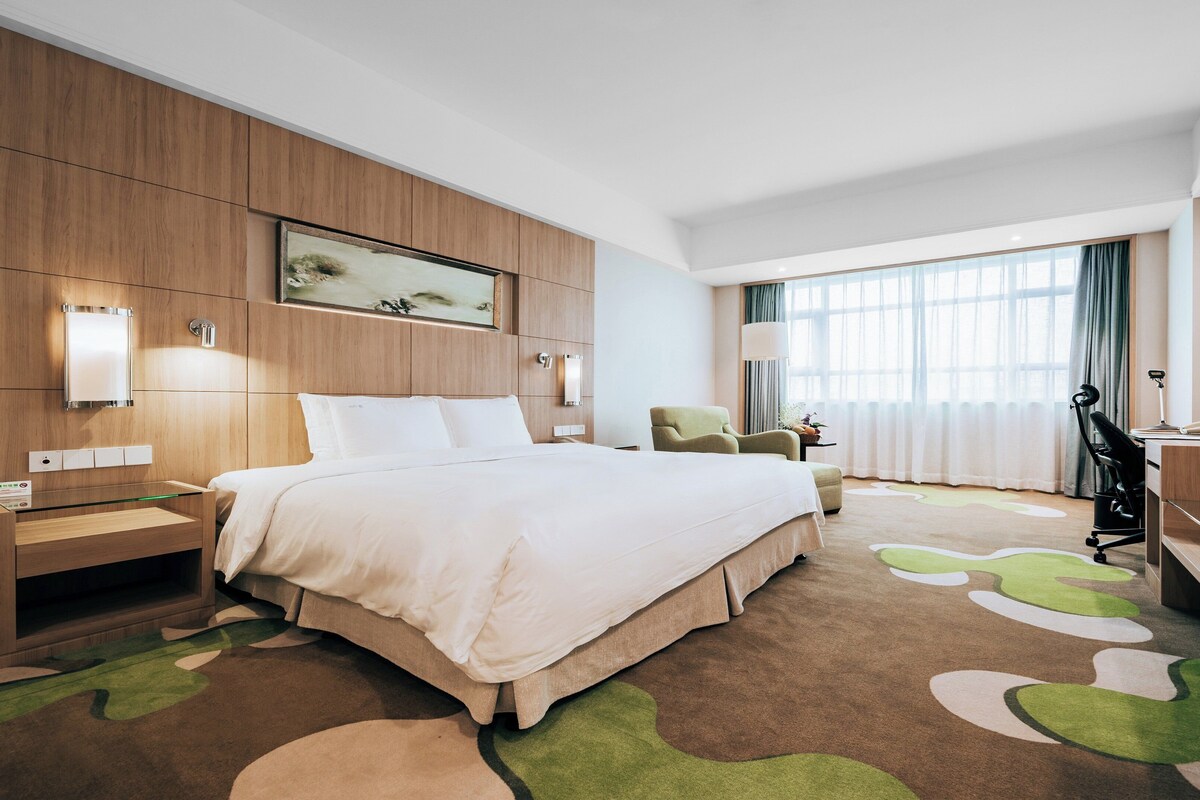 A hotel room with a large bed, a desk, a chair, a desk lamp, and a window with curtains.