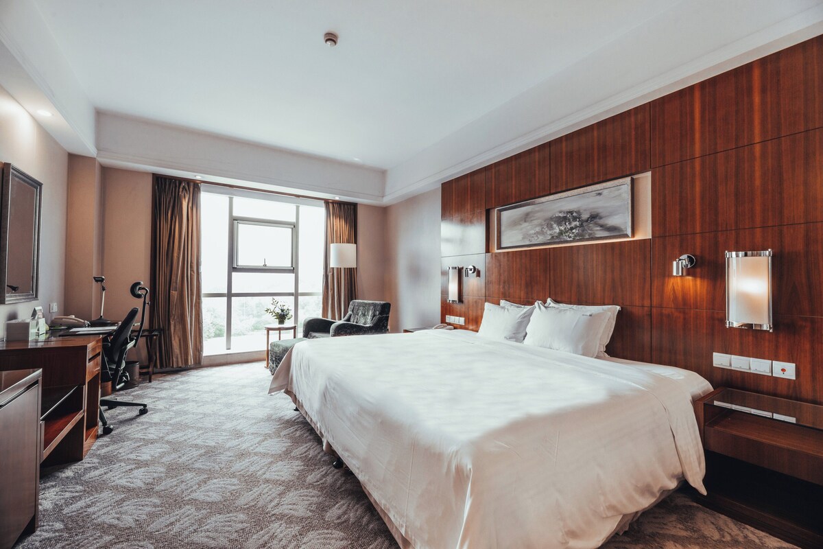 A hotel room with a large bed, a desk, a chair, a lamp, and a window with curtains.