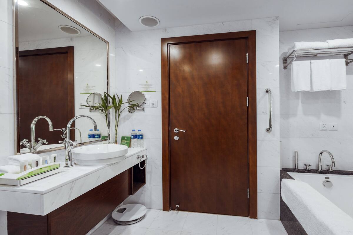 A modern bathroom with a wooden door, a large mirror, a sink with a scale, a bathtub, and a towel rack.