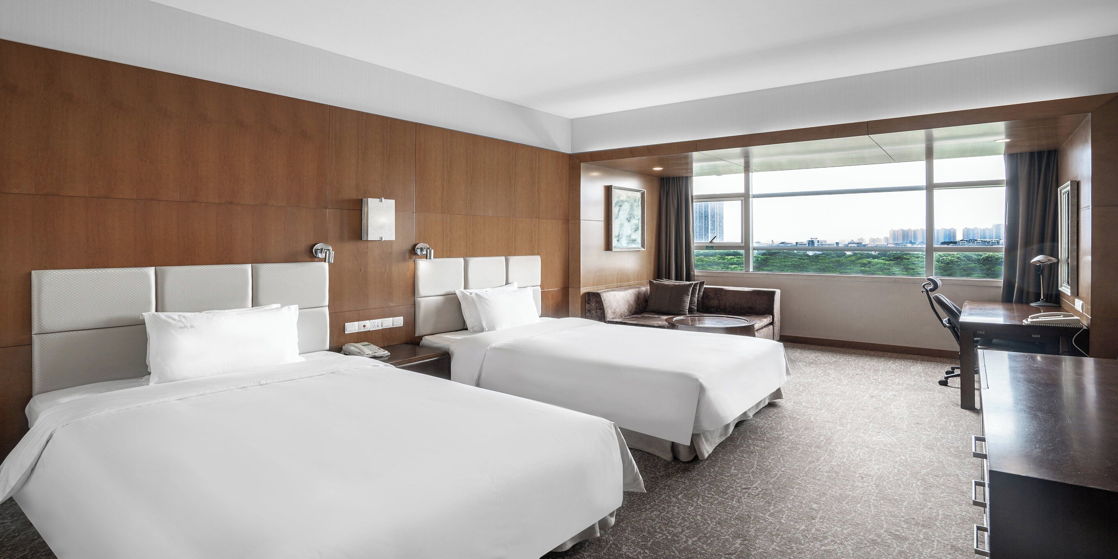 Habitación de hotel con dos camas, un escritorio, una silla, ventana con vista a la ciudad y un espejo grande.