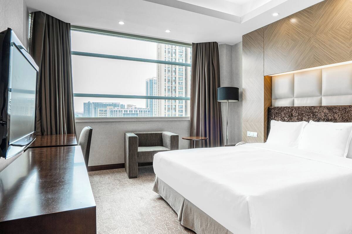 A modern hotel room with a large bed, a desk, a chair, a TV, and a window with a city view.