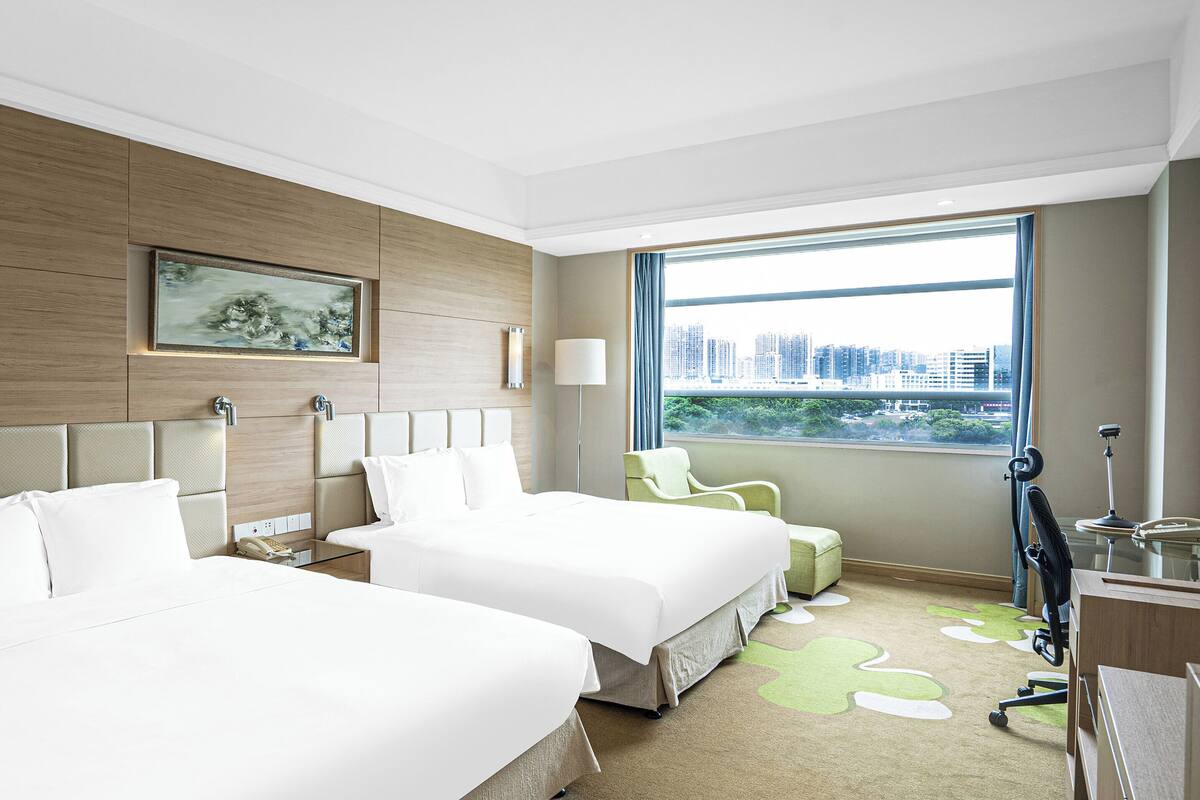 A hotel room with two beds, a desk, a chair, and a large window with a city view.