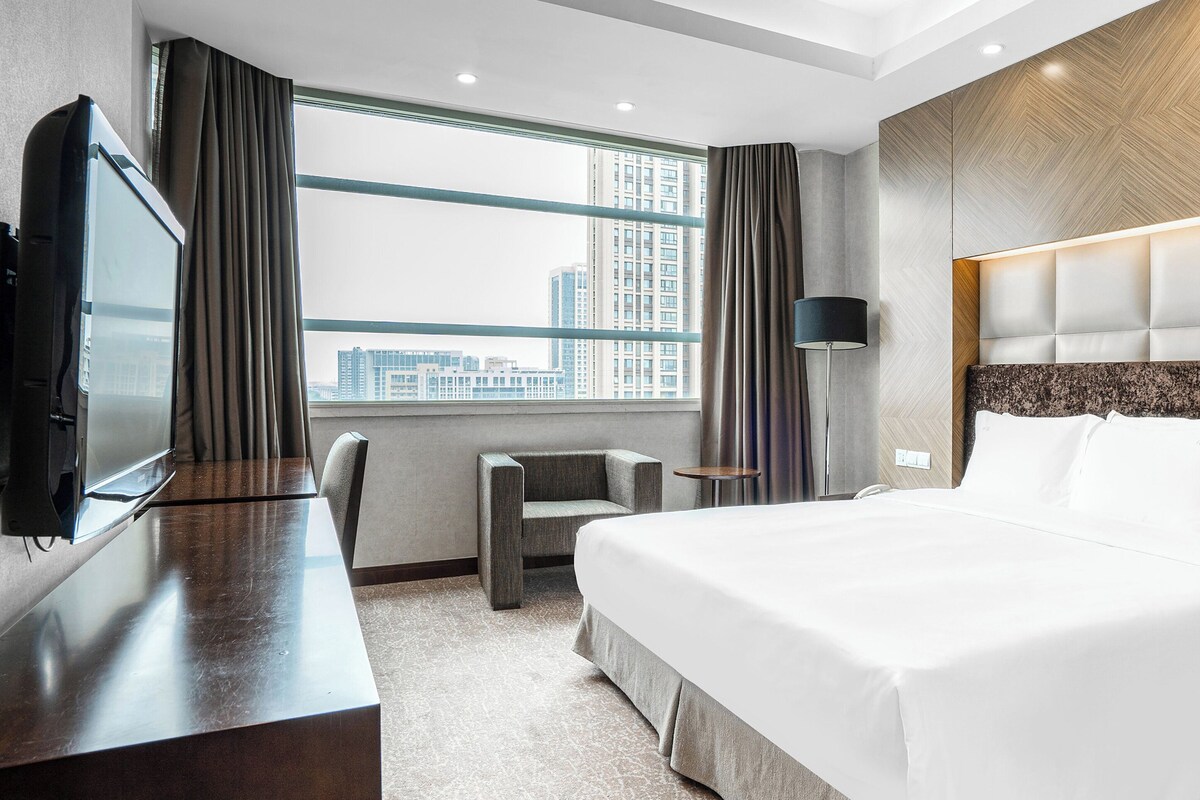 A modern hotel room with a large bed, a desk, a chair, a TV, and a window with a city view.