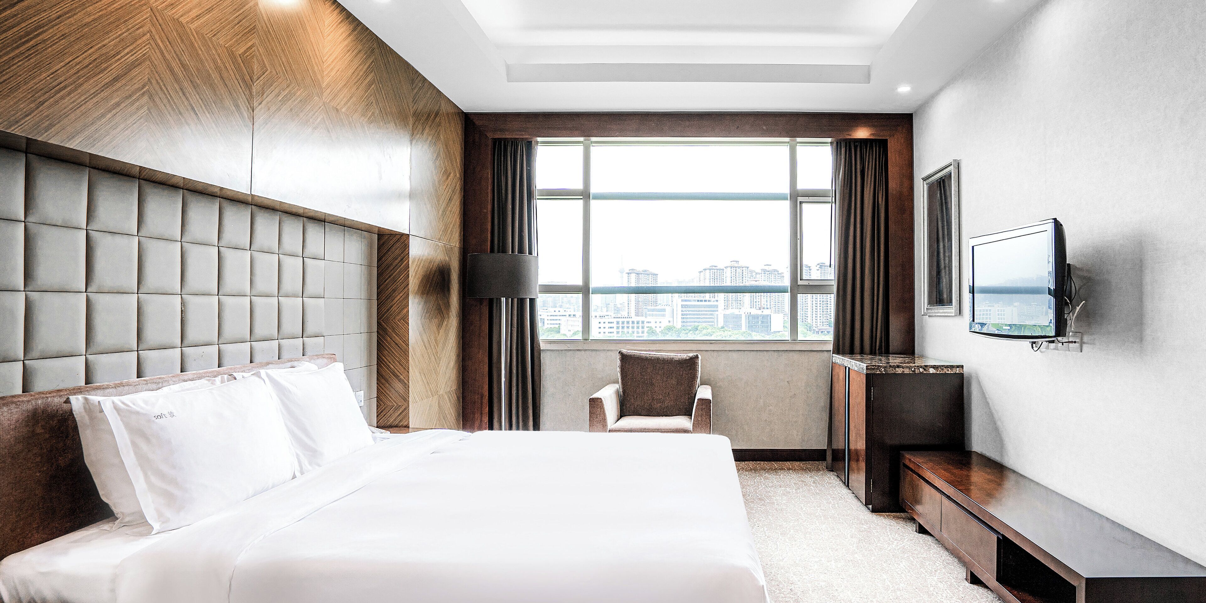 Ein Hotelzimmer mit einem großen Bett, einem Fernseher, einem Sessel und Blick auf die Stadt.