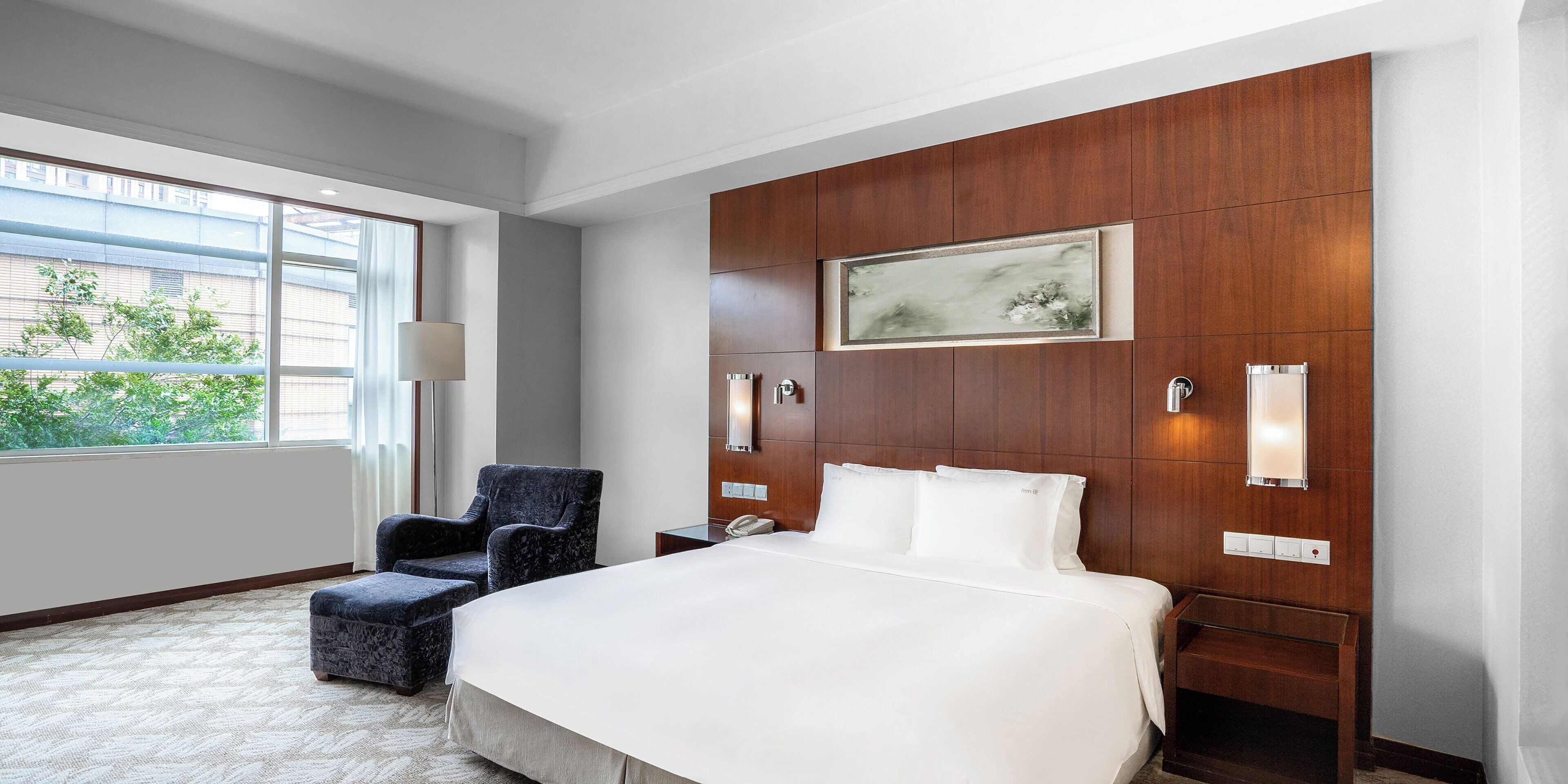 Habitación de hotel con una cama grande, una silla, una mesita de noche, una lámpara y una ventana con vistas a edificios y árboles.