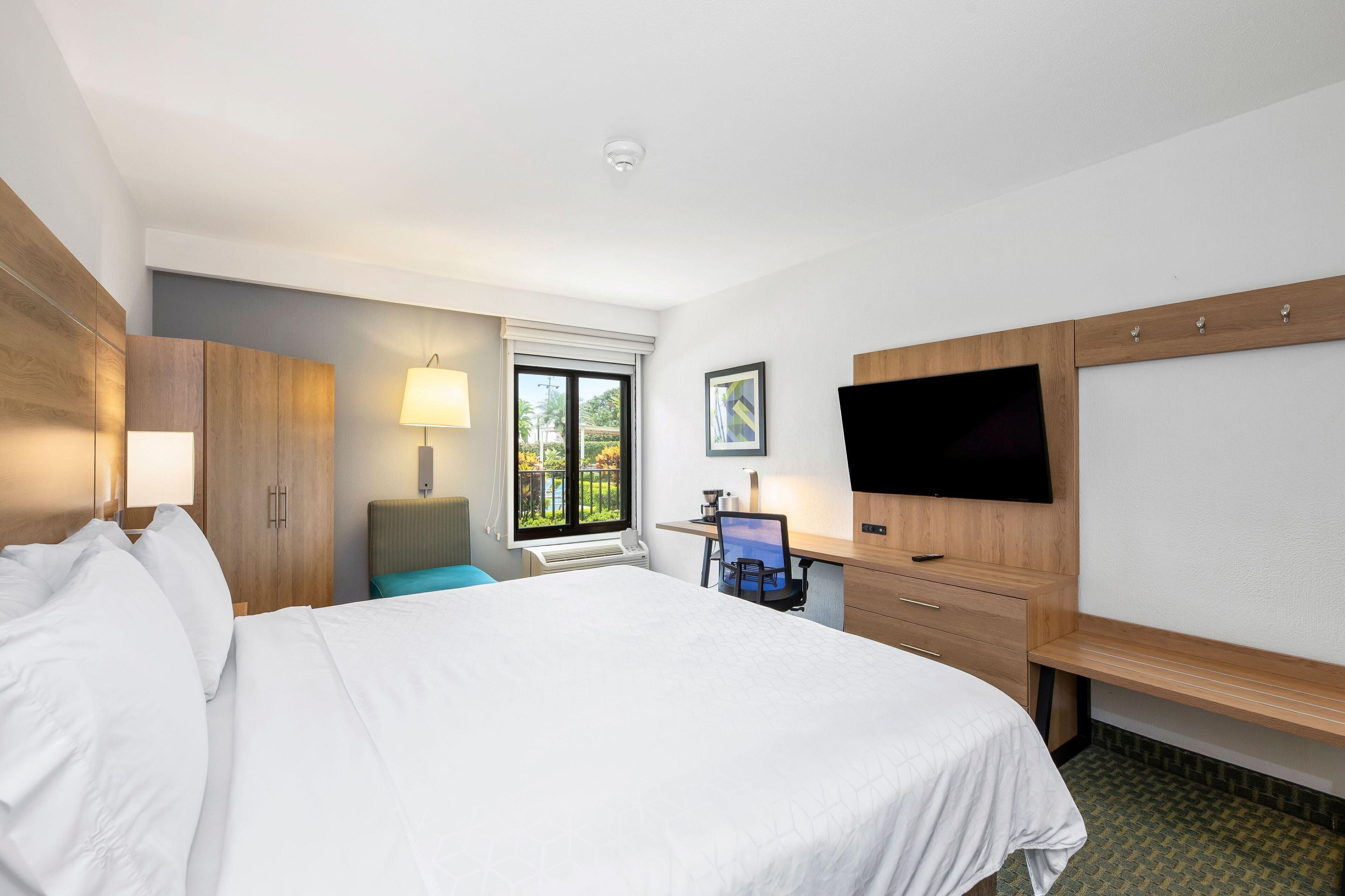 A hotel room with a large bed, a desk with a computer, a TV, and a window with a view of greenery.