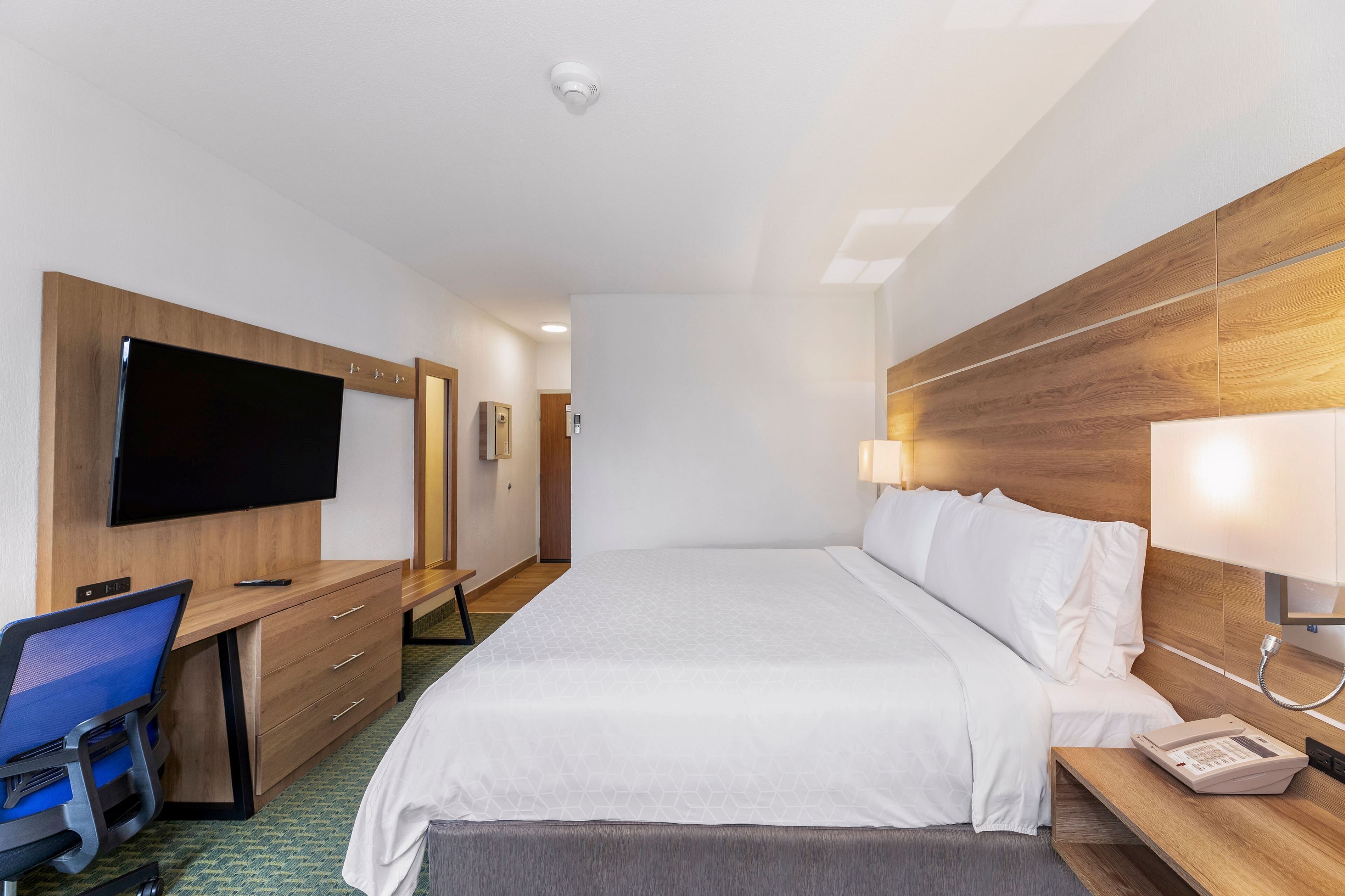 A hotel room with a bed, a desk with a chair, a television, and a bedside table with a lamp and phone.