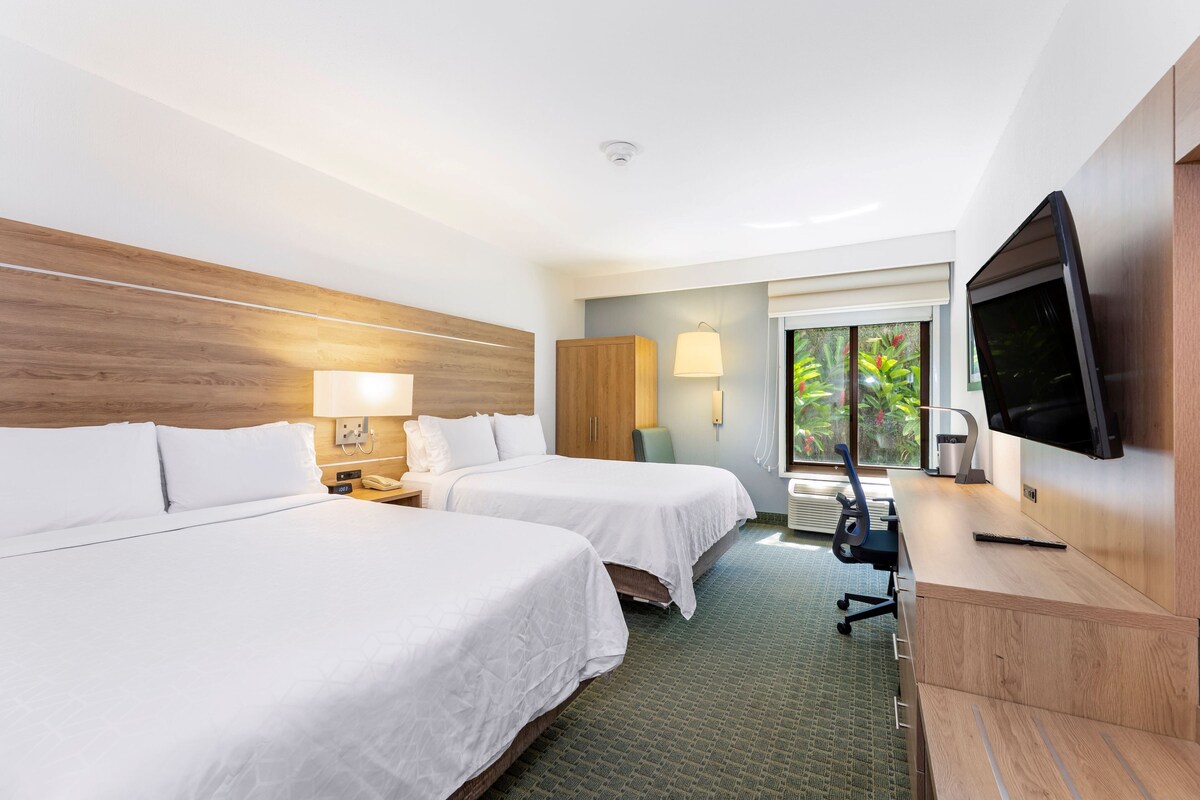 A hotel room with two beds, a desk, a television, and a window with a view of greenery.
