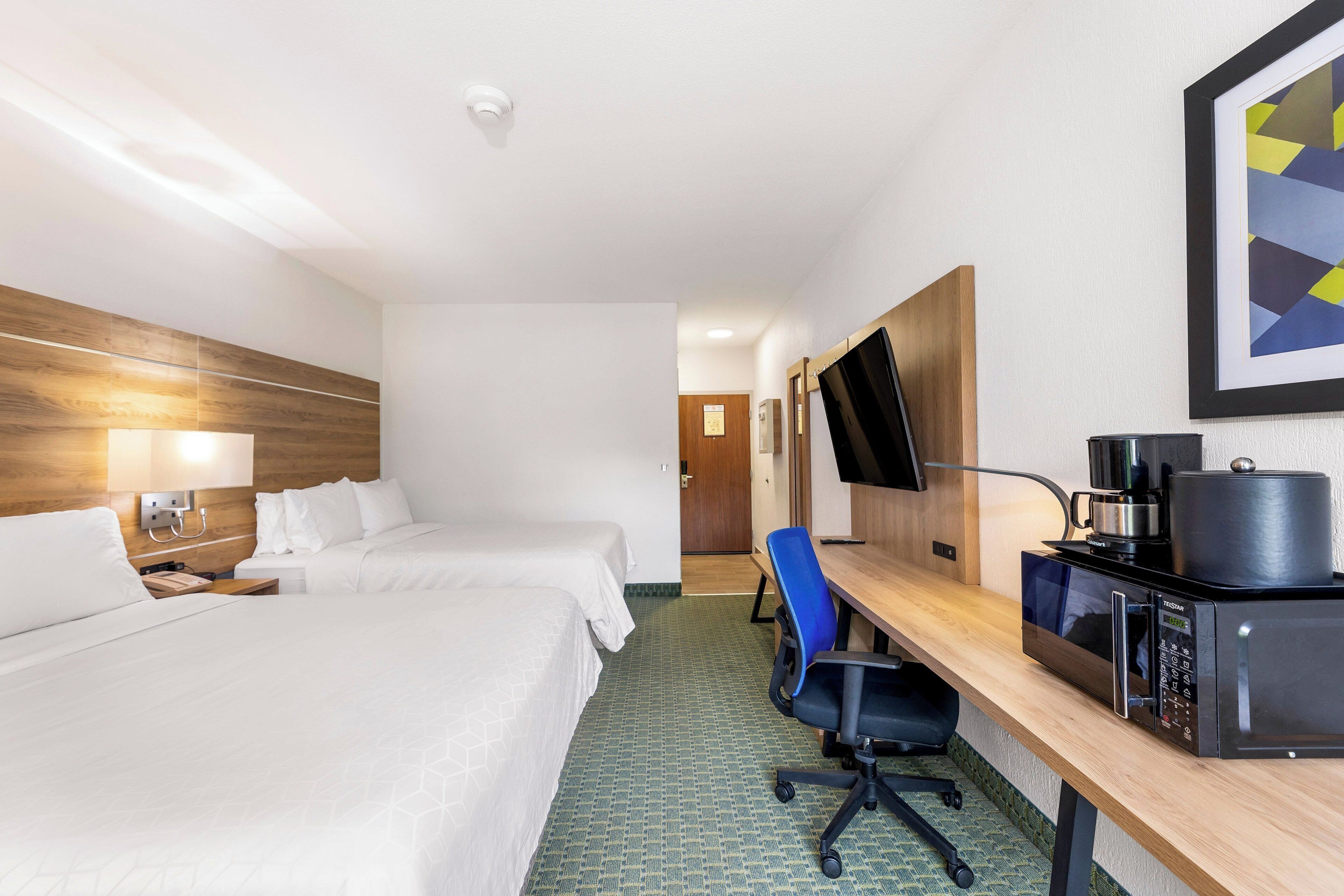 A hotel room with two beds, a desk with a microwave, a chair, and a television on the wall.