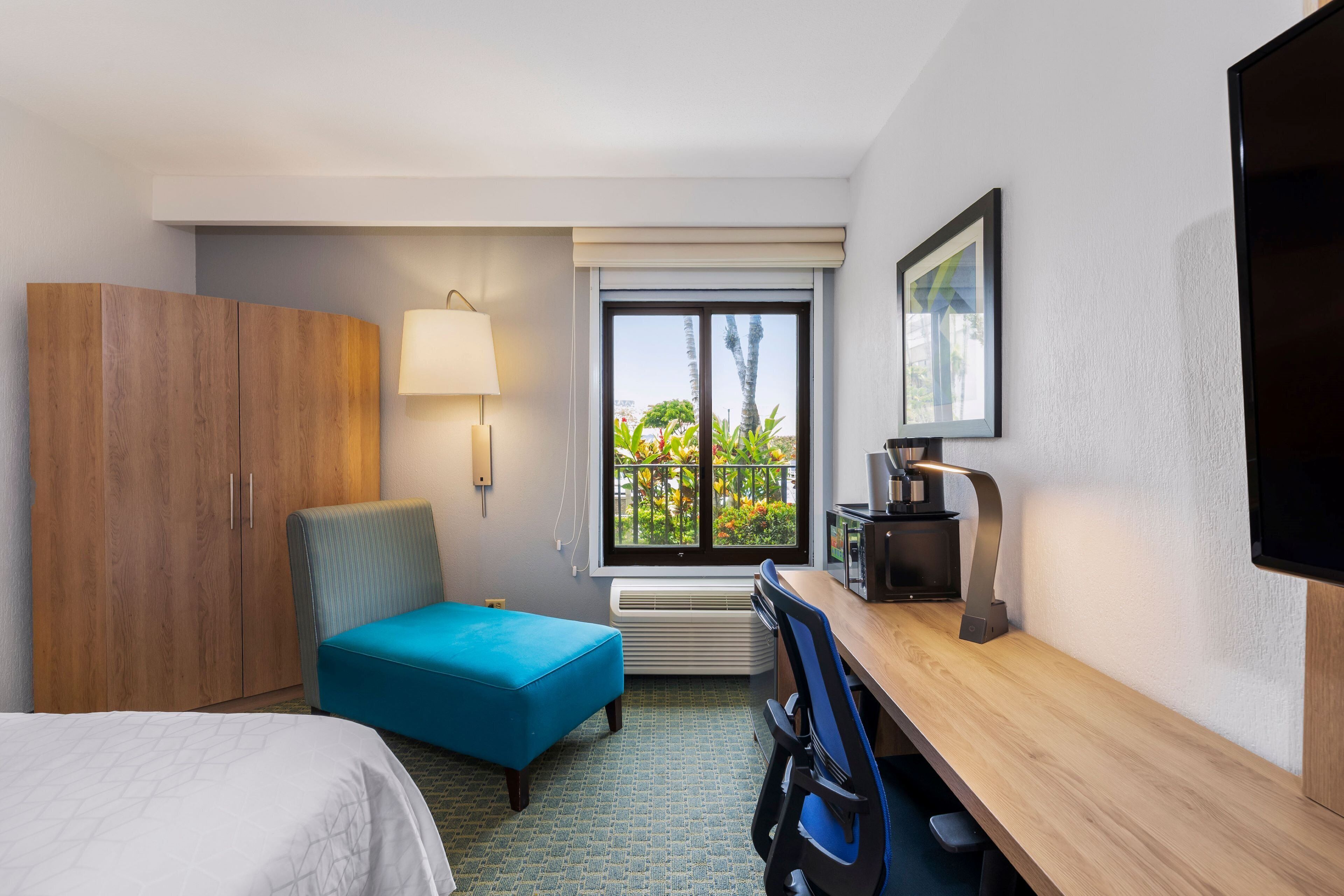 A hotel room with a bed, a desk with a chair, a blue ottoman, a wardrobe, a window with a view of greenery, and a flat-screen TV.