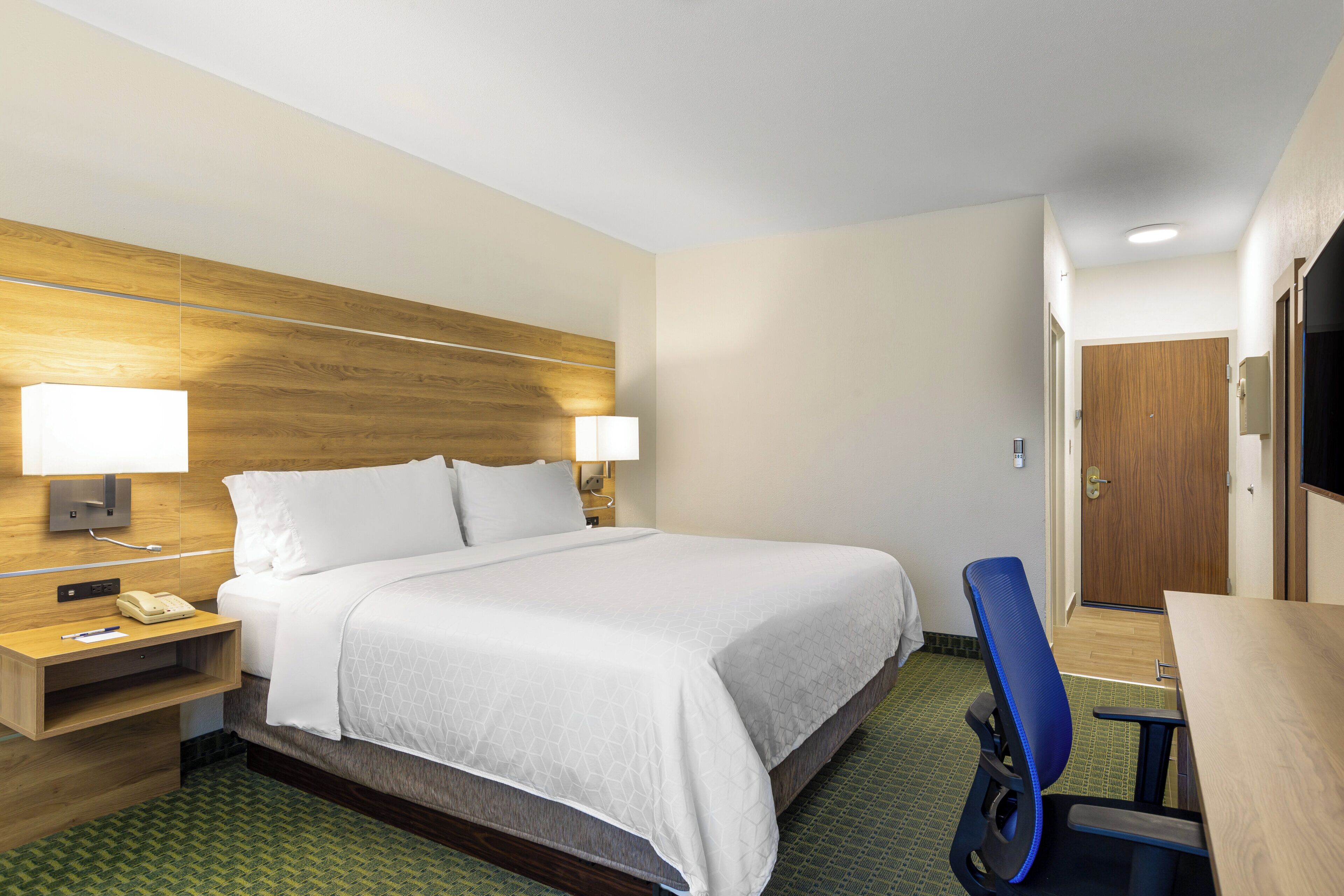 Habitación de hotel con cama, mesita de noche, lámpara, teléfono y escritorio con silla.