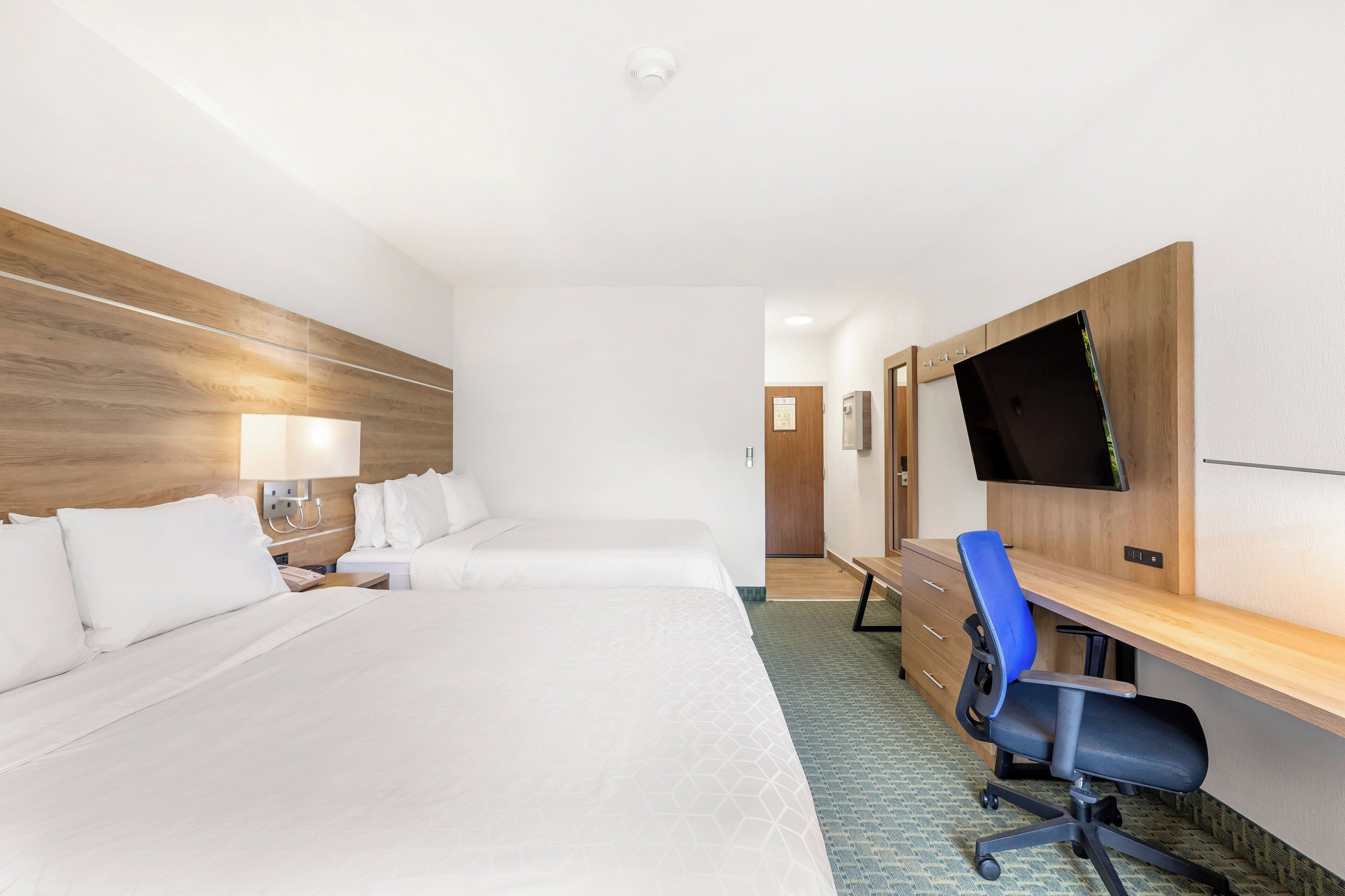 A hotel room with two beds, a desk with a chair, a flat-screen TV, and a wooden headboard.