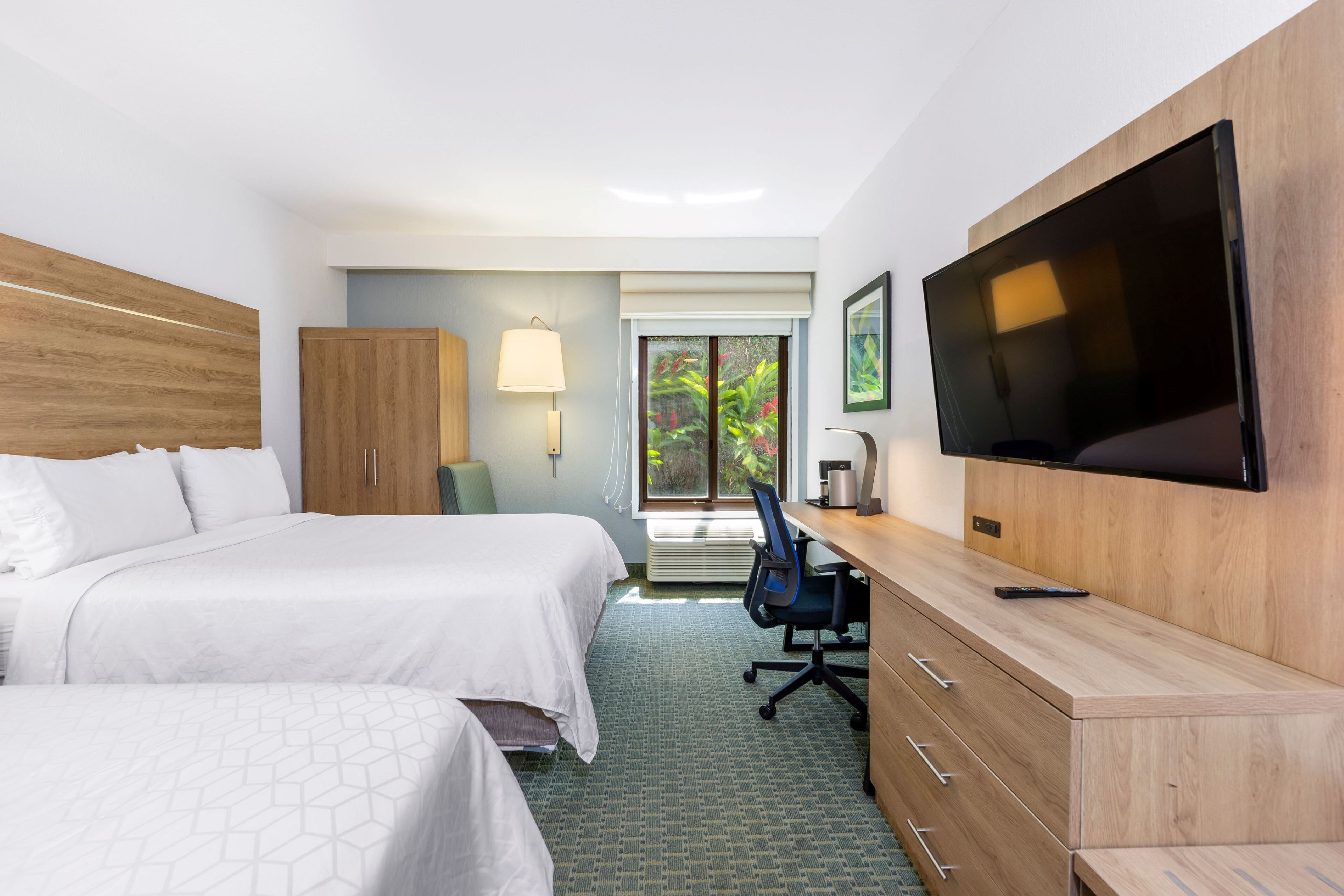 A hotel room with two beds, a desk, a TV, and a view of greenery outside.