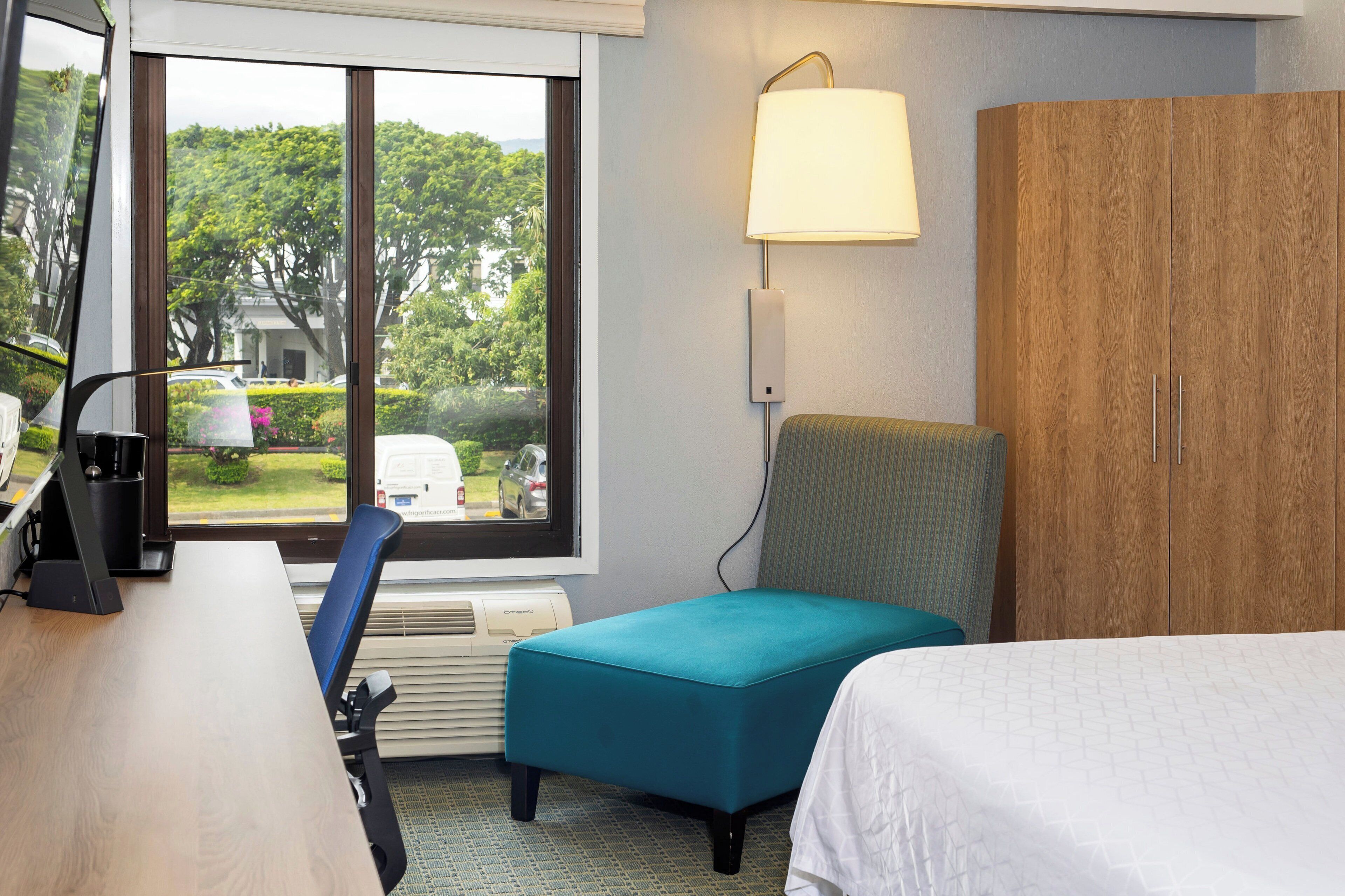 Ein Hotelzimmer mit Schreibtisch, blaugrünem Ottomanen, einem Bett, einem Holzschrank und einem Fenster mit Blick auf Grünflächen und parkende Autos.