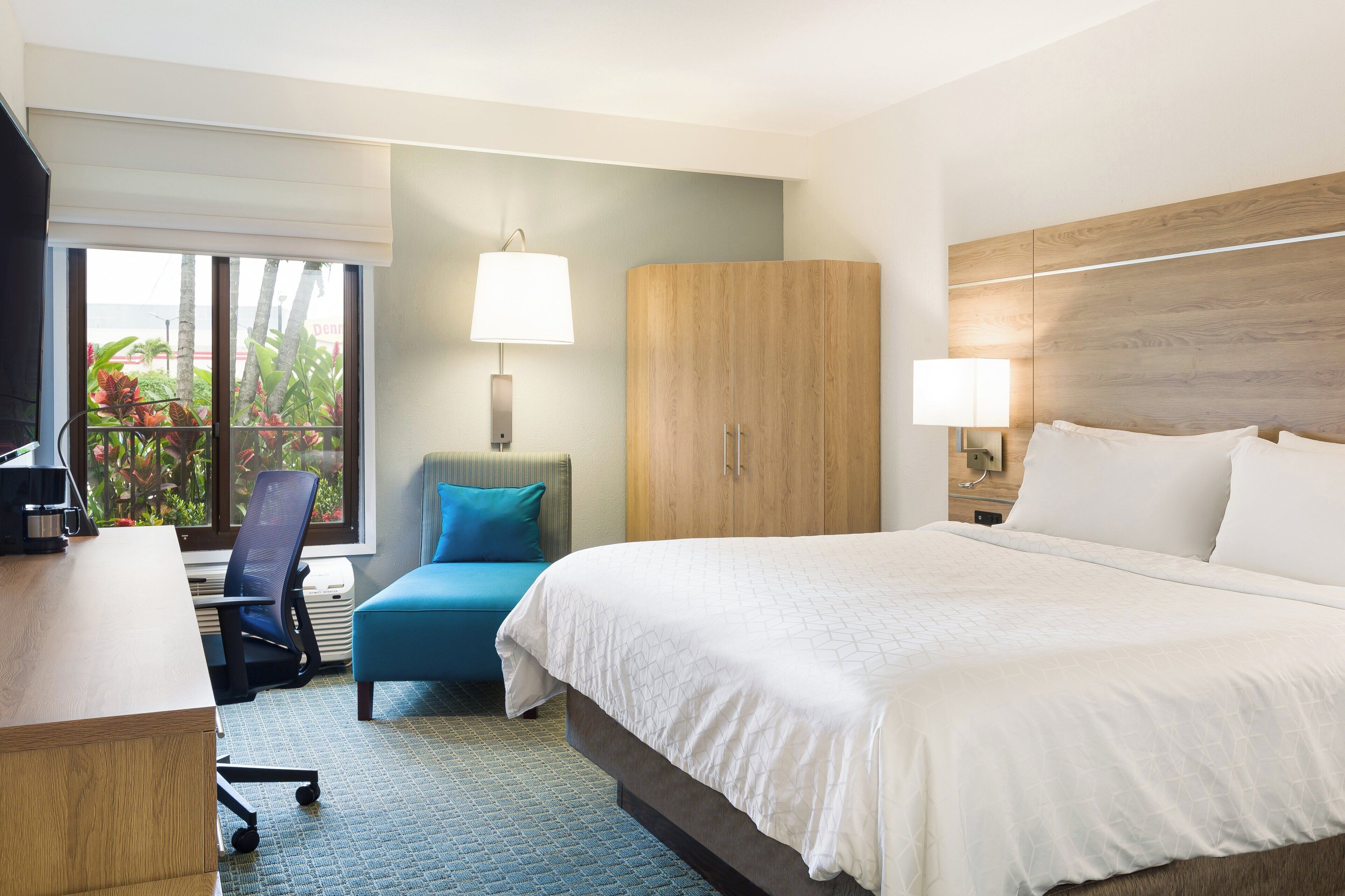 A hotel room with a bed, a desk, a chair, a wardrobe, and a window with a view of greenery.