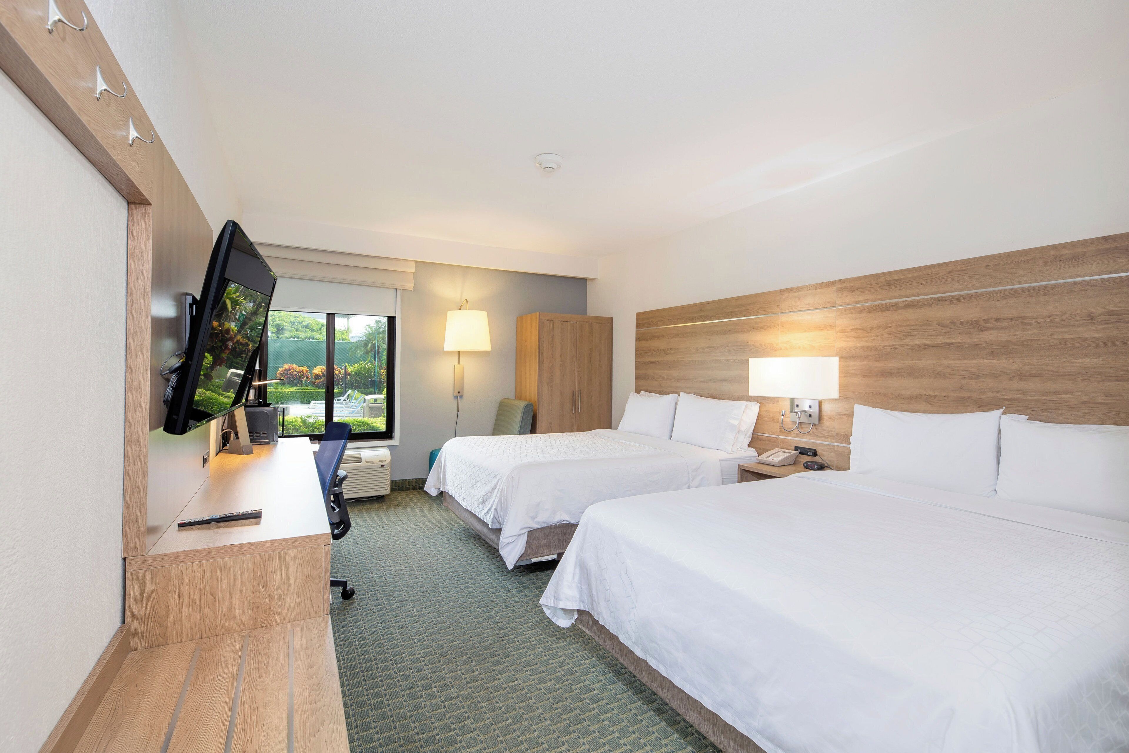 A hotel room with two beds, a desk with a computer, a TV, and a window with a view of greenery.